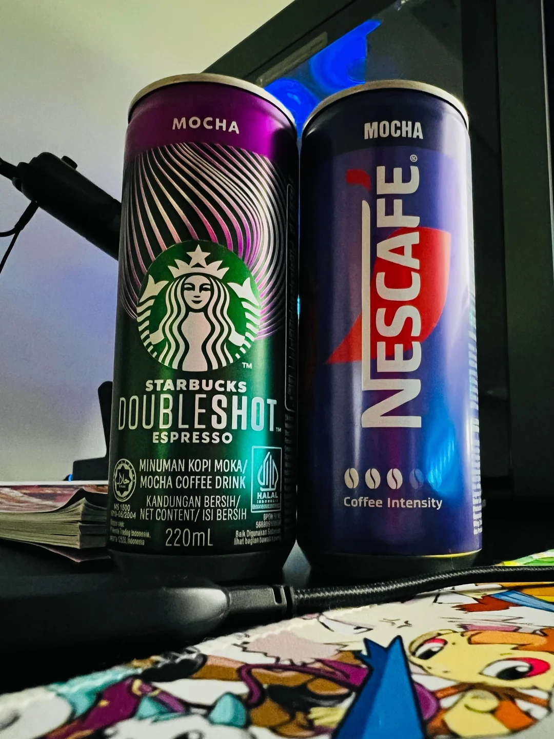 Two cans of mocha coffee drinks—Starbucks Doubleshot Espresso and Nescafe—are placed side by side on a desk, with part of a colorful cartoon-printed surface and a computer monitor visible in the background.