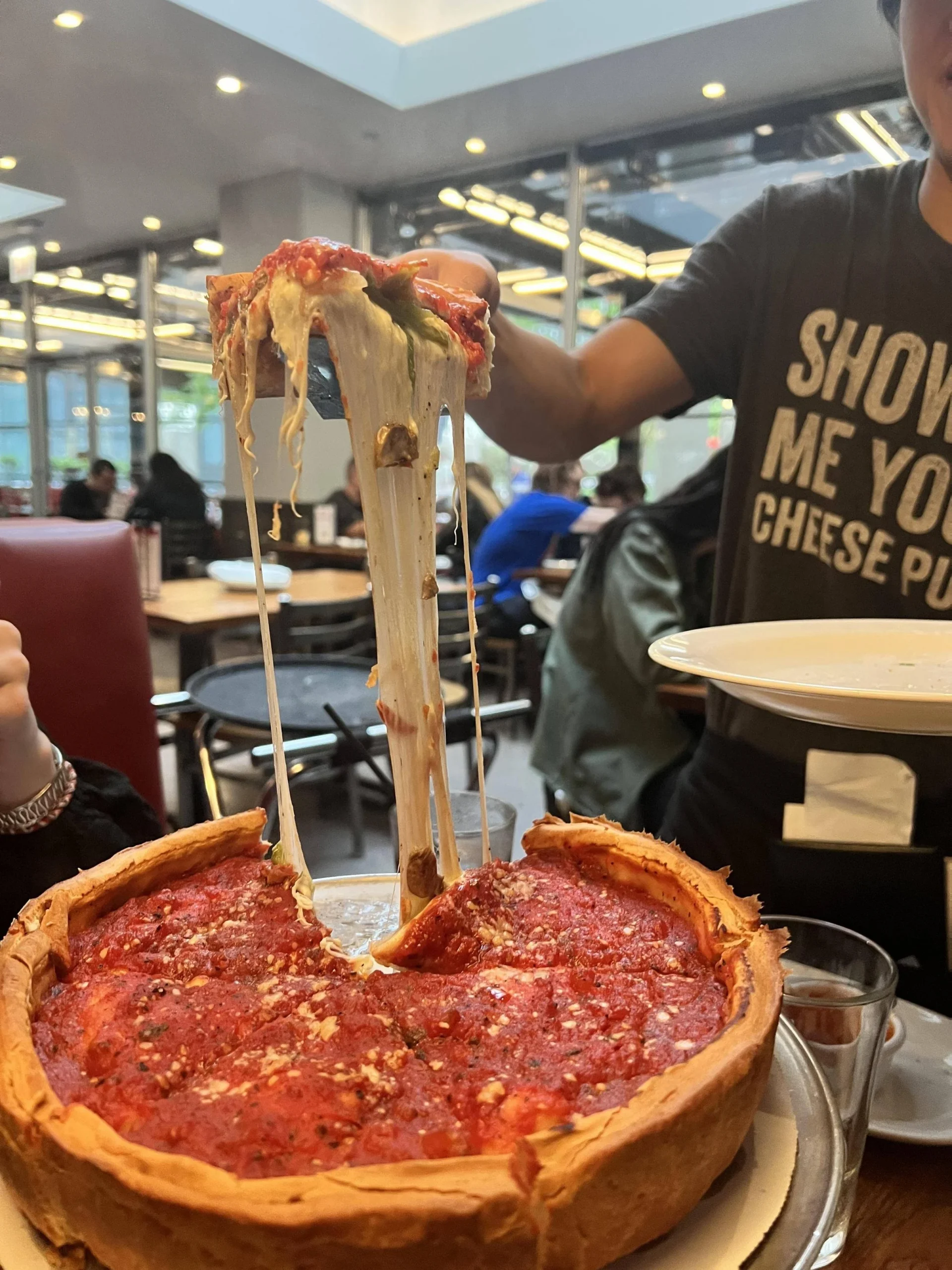 A person lifts a gooey slice of deep-dish pizza with melted cheese stretching from the pan. The pizza is thick with tomato sauce and cheese. Indoor dining tables and a person wearing a "SHOW ME YOUR CHEESE PULL" shirt are visible.