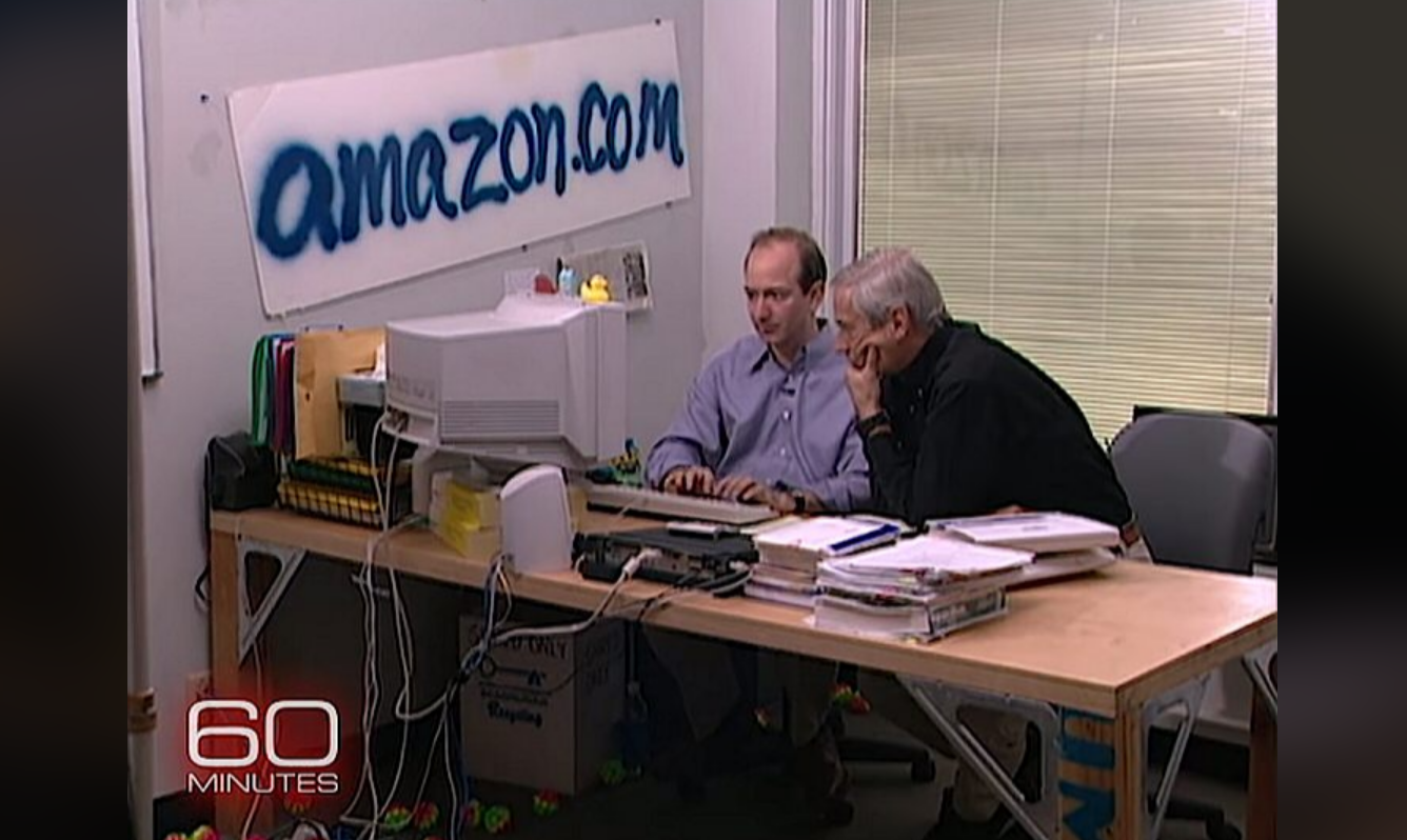 Two men sit at a cluttered desk with a computer in a simple office. A hand-painted amazon.com sign is on the wall. Papers and office supplies cover the table. The "60 Minutes" logo appears in the bottom left corner.