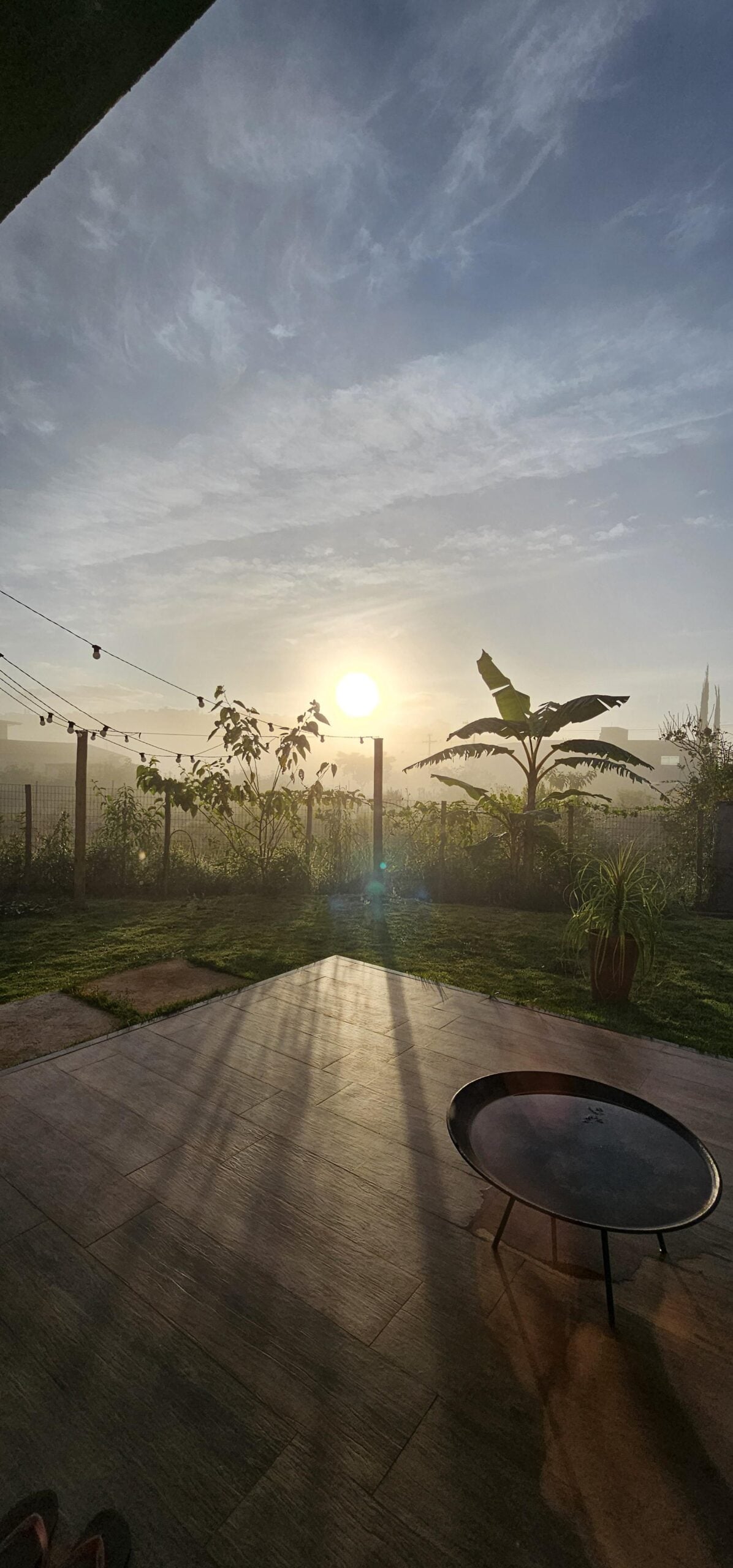 Sunrise over a lush backyard garden with green plants, banana trees, and string lights. The sun casts long shadows onto a tiled patio, where a black round fire pit sits in the foreground.