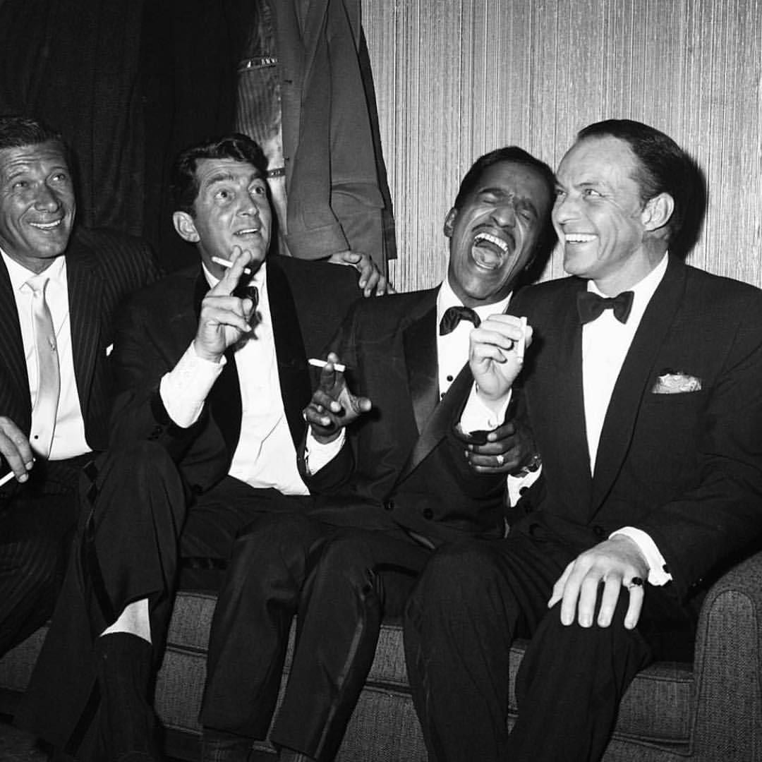 Four men in tuxedos sit closely together on a couch, laughing and holding cigarettes, clearly enjoying each other's company in a lively, sophisticated setting.