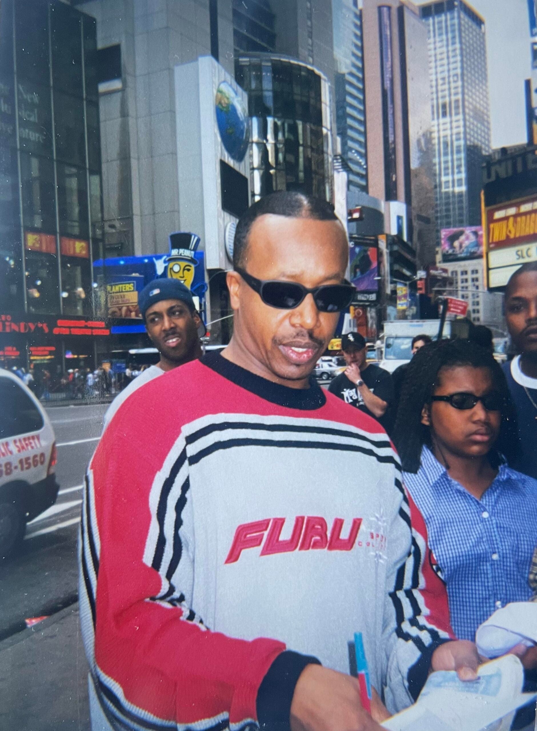 A man wearing sunglasses and a red, white, and gray FUBU sweater signs an autograph on a busy city street, with several people and tall buildings in the background.