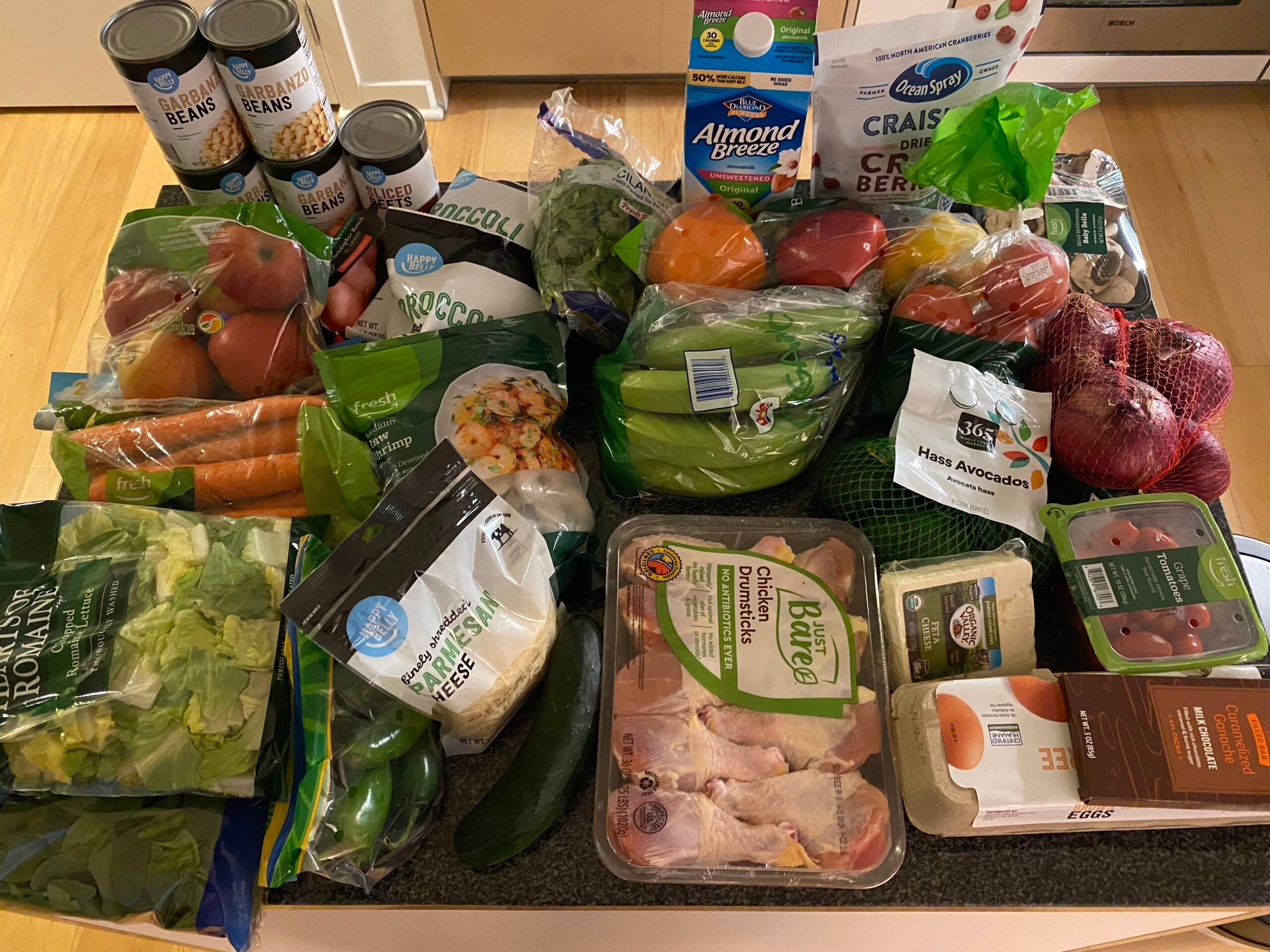 A variety of groceries on a counter, including fresh vegetables, canned beans, chicken, eggs, almond milk, salad greens, bananas, avocados, tomatoes, and a bag of dried cranberries.