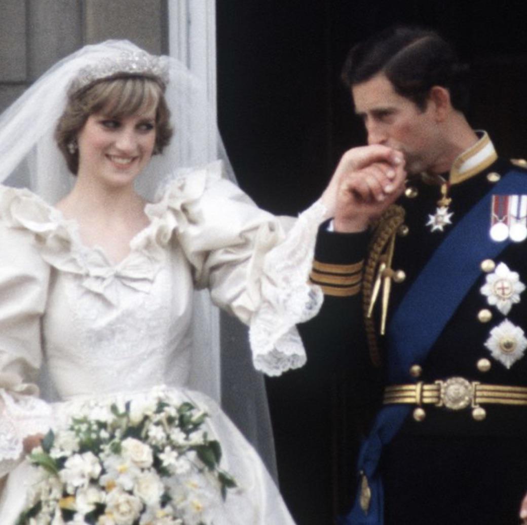 A bride in a white wedding dress and veil holds a bouquet of flowers while a groom in a decorated military uniform kisses her hand. They are smiling and standing close together.