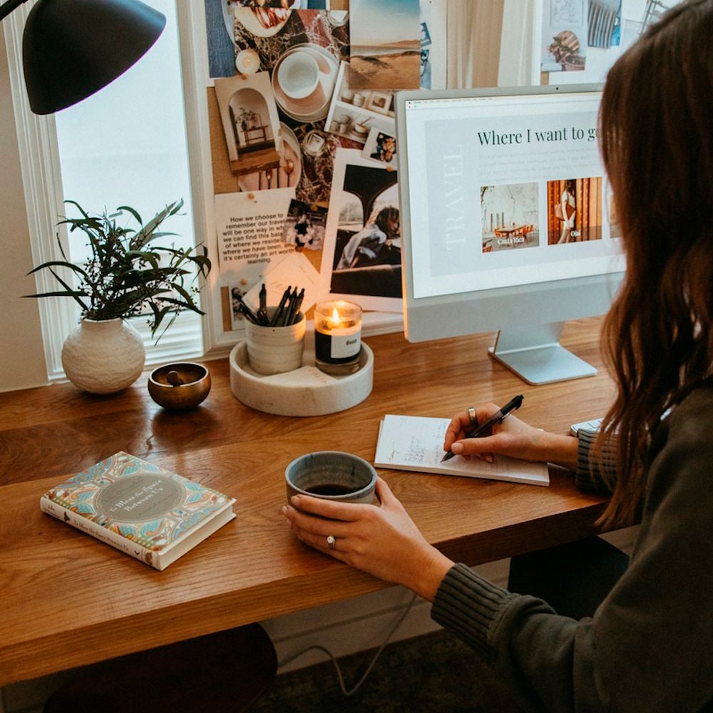 A woman sits at a wooden desk, holding a mug and writing in a notebook. An open journal, candle, potted plant, and computer displaying travel plans are on the desk. A photo collage decorates the wall.