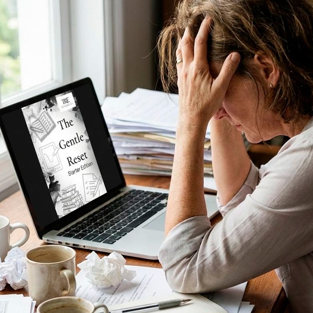 A woman sits at a cluttered desk with her head in her hands, looking stressed. An open laptop displays an e-book titled "The Gentle Reset" on the screen. Crumpled papers and empty mugs are on the desk.