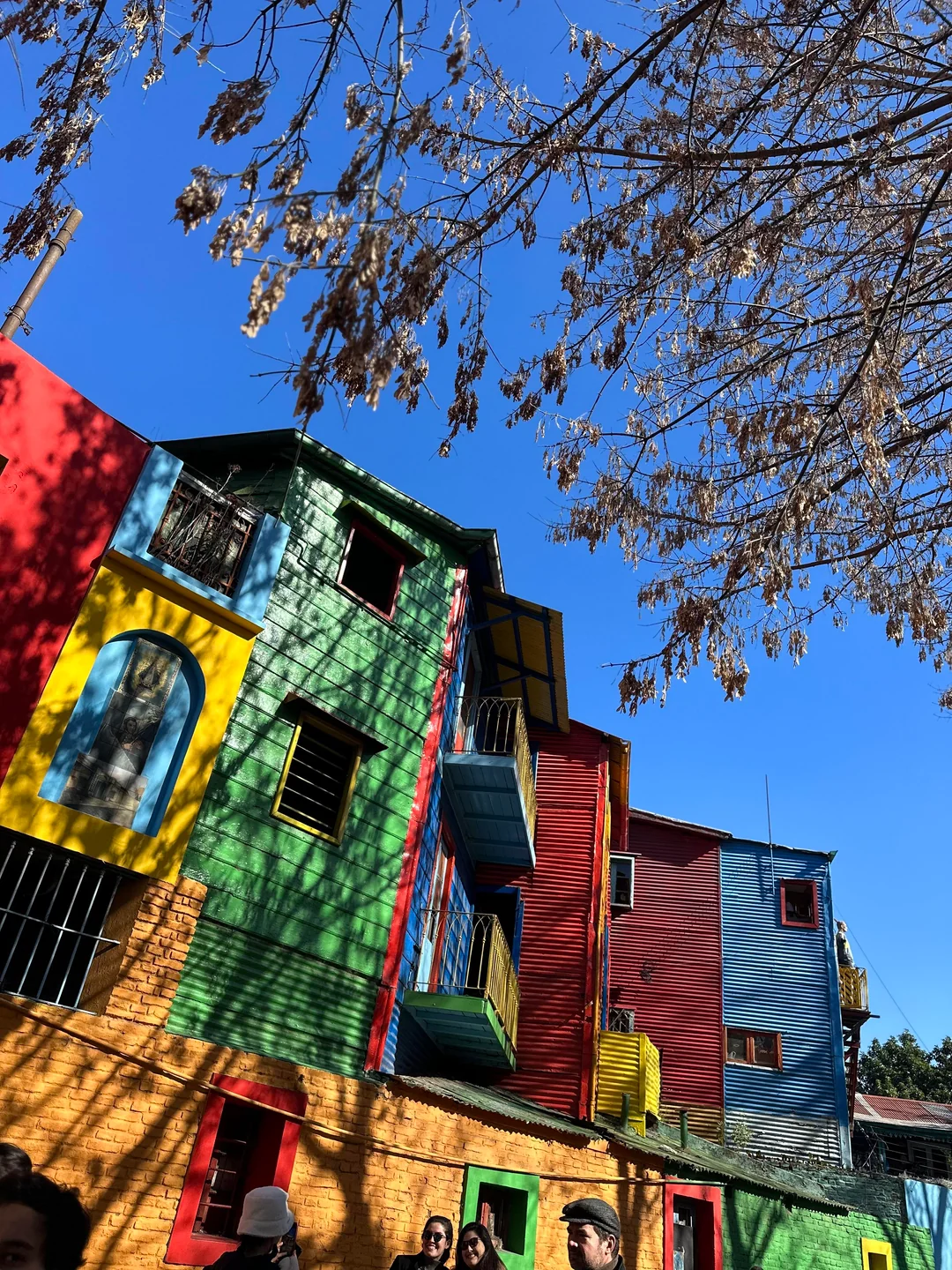 Colorful houses with red, green, yellow, and blue facades stand side by side under a clear blue sky. Leafless tree branches cast shadows on the buildings, and a few people are visible at the bottom of the image.