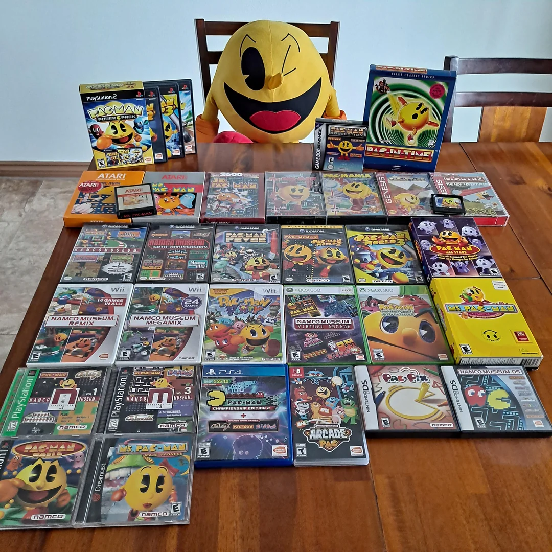 A large collection of Pac-Man games, memorabilia, and collectibles is neatly arranged on a wooden table. A big Pac-Man plush sits on a chair behind the table, surrounded by various game boxes and discs from different consoles.