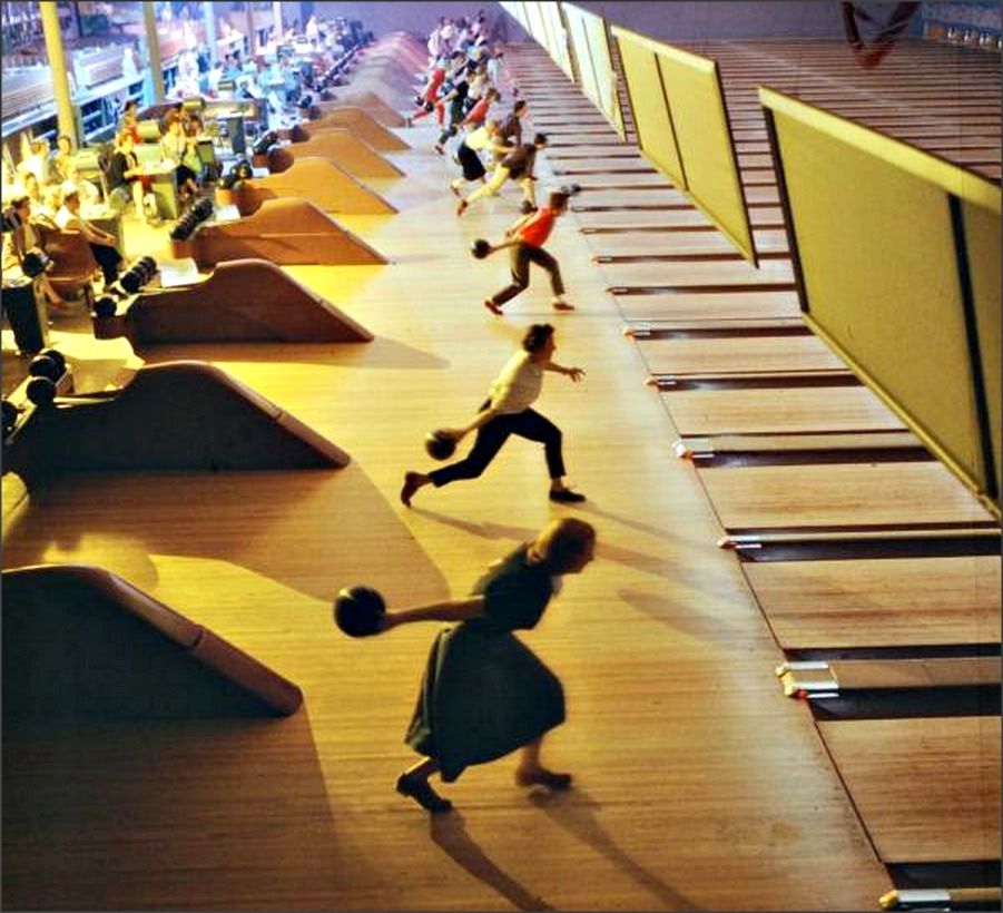 A group of people bowling at a busy bowling alley, each on their own lane, captured mid-throw as they release their bowling balls toward the pins. The scene is lively and full of motion.