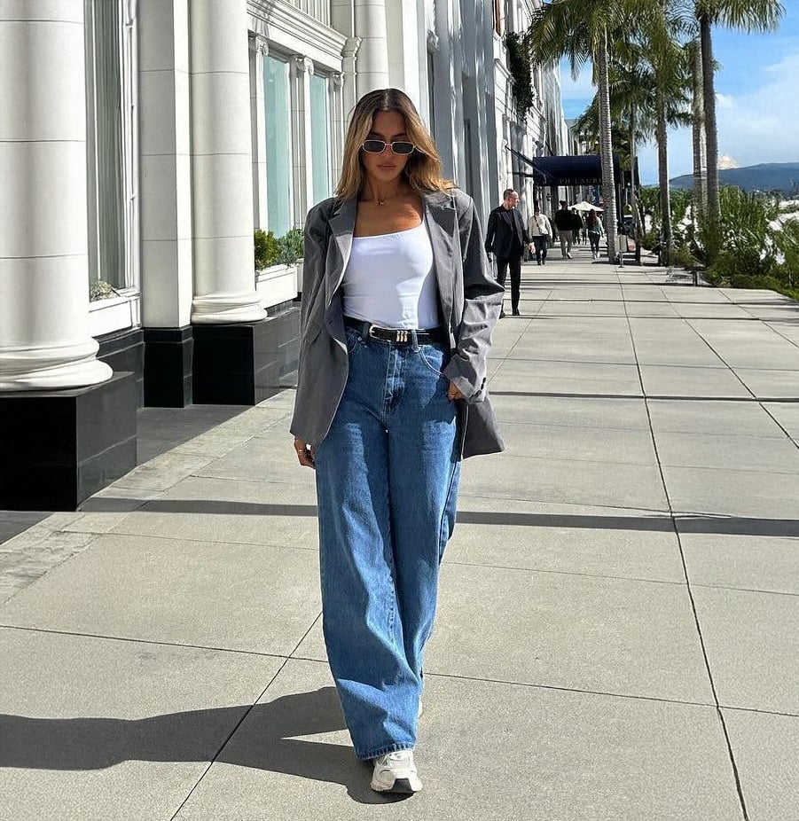 A woman wearing sunglasses, a gray blazer, white top, blue wide-leg jeans, and white sneakers walks confidently on a sunny city sidewalk lined with palm trees and white columns.
