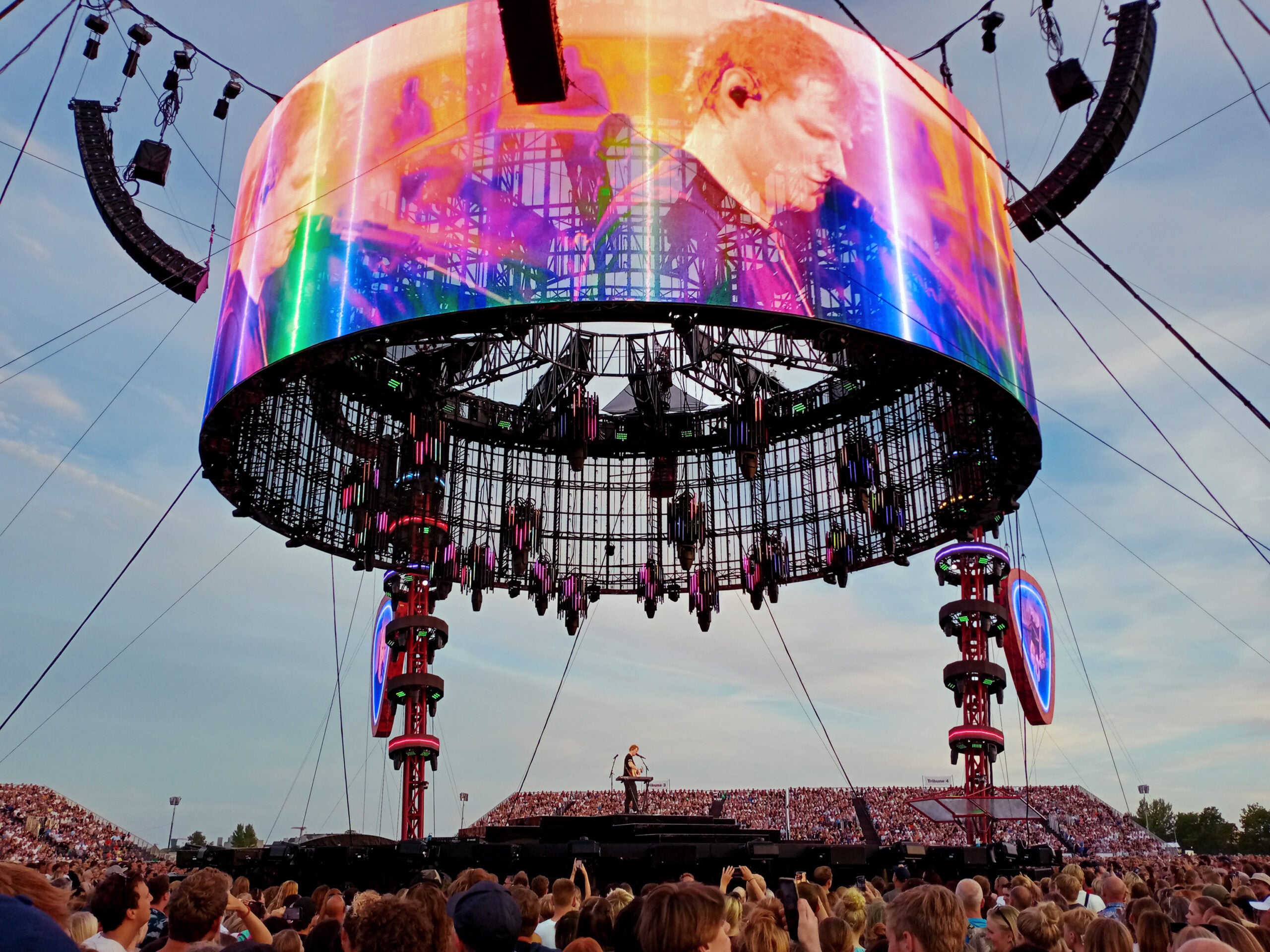 A musician performs on a large outdoor stage under a circular screen displaying his image, surrounded by a crowd of people at sunset.