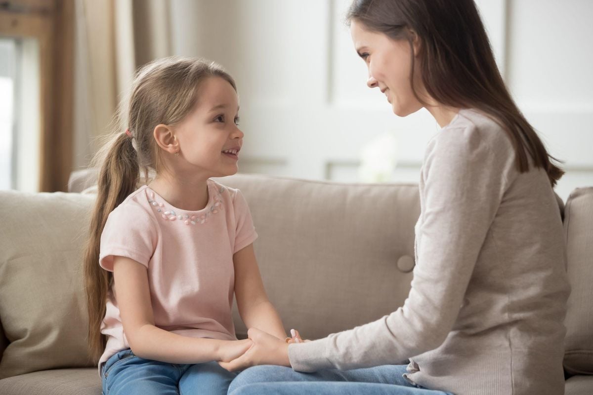 A woman and a young girl sit on a beige couch, facing each other and smiling warmly while holding hands, suggesting a close and caring conversation.