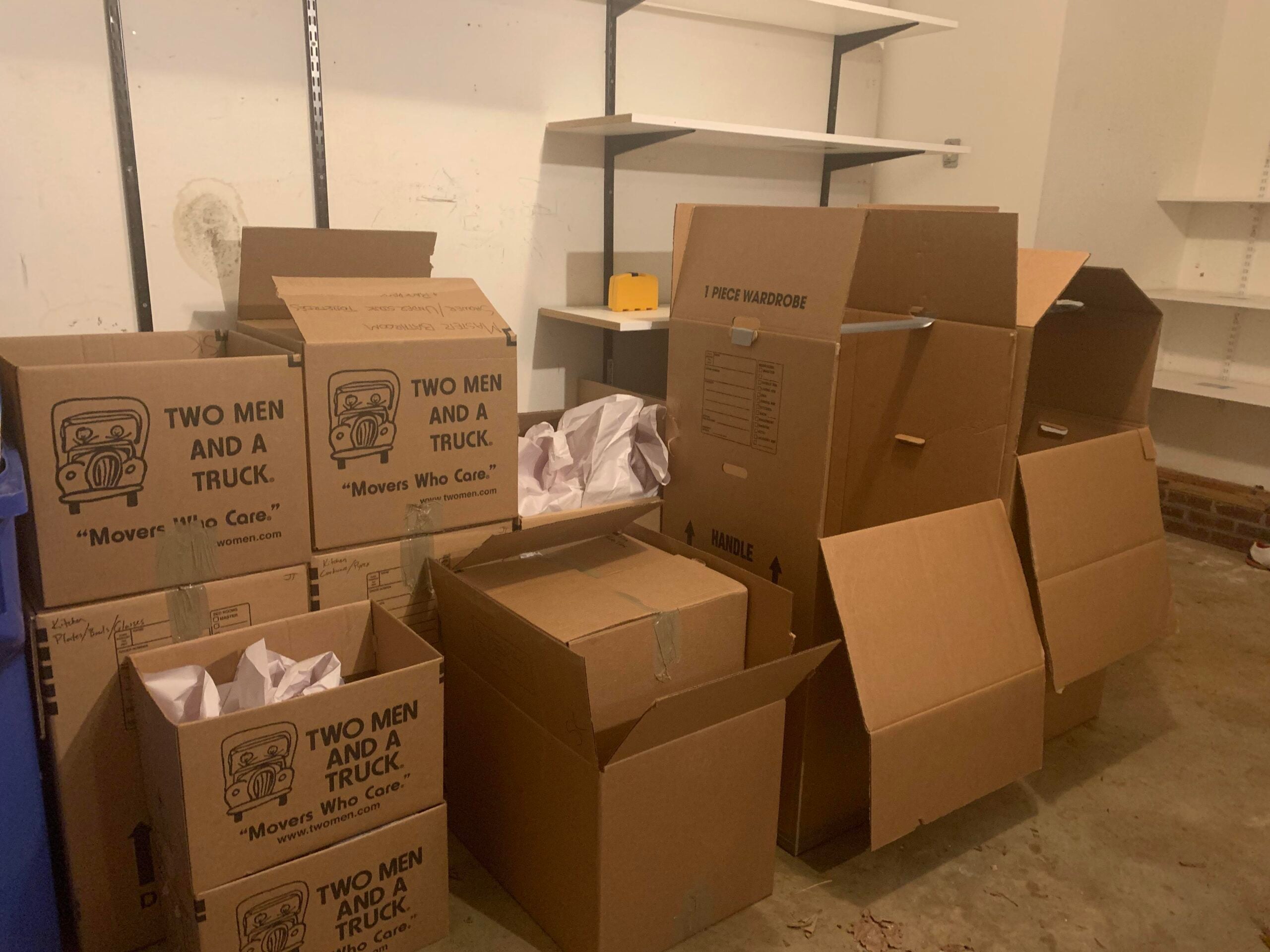 Several cardboard moving boxes, some labeled "Two Men and a Truck," are stacked and open in a mostly empty room with shelves and a concrete floor. Packing paper is visible in some boxes.