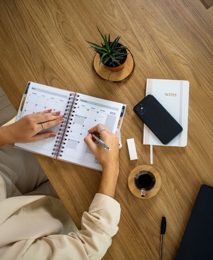 A person writes in a spiral planner on a wooden desk with a small potted plant, a notebook, a smartphone, a cup of coffee, a pen, and an eraser nearby.