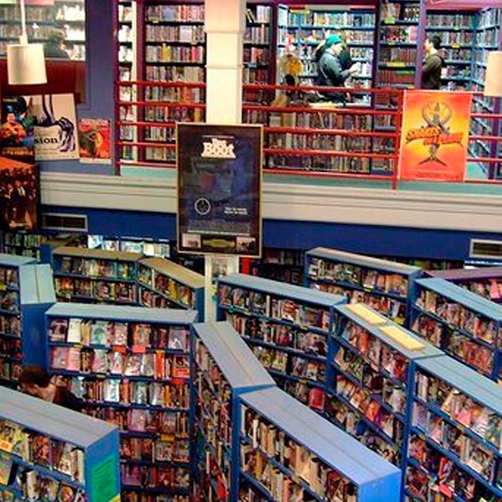 A colorful library or bookstore with blue bookshelves filled with books, two floors visible, people browsing upstairs, and bright posters on the walls.