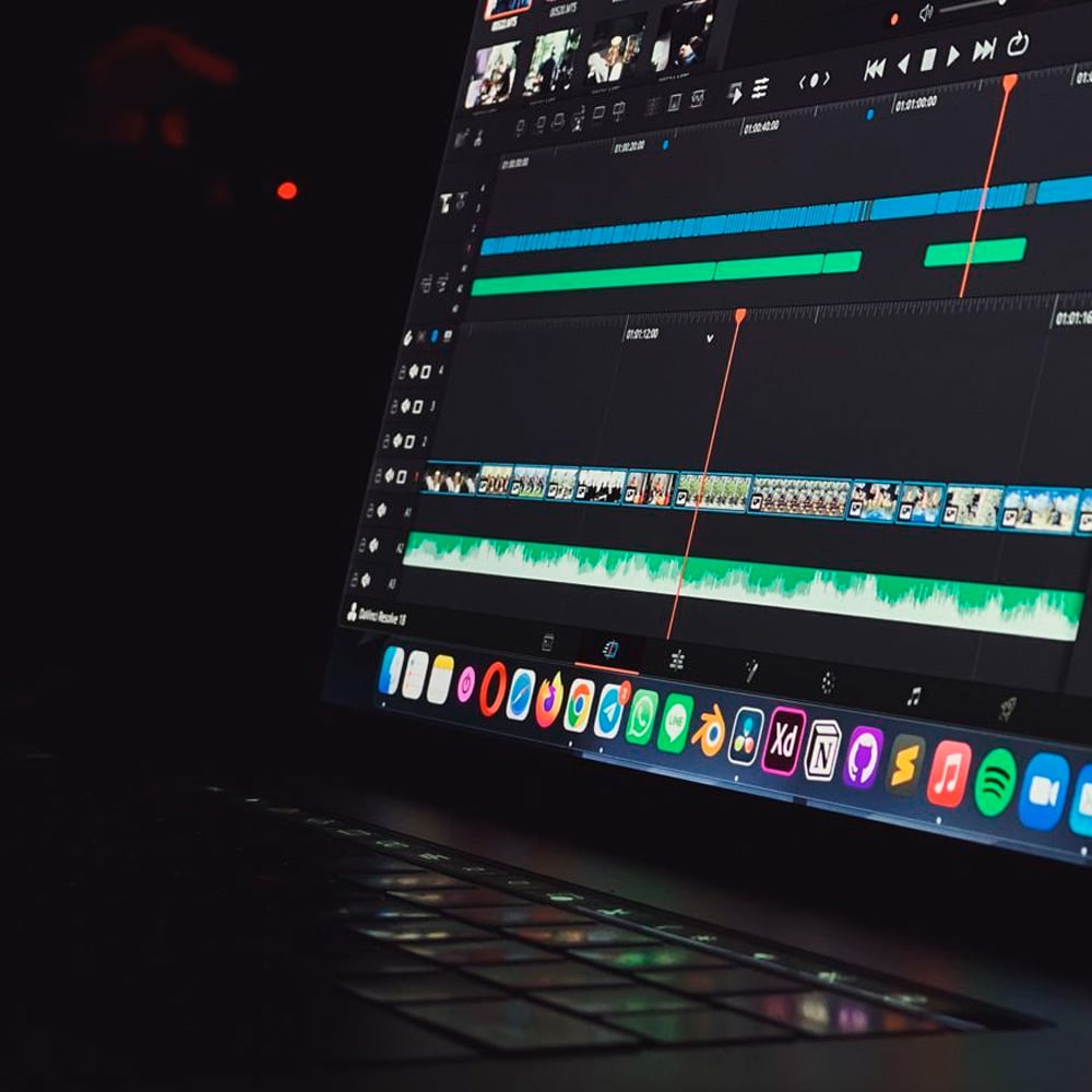 A close-up of a laptop screen displaying a video editing software interface with timelines, audio tracks, and various editing tools; colorful application icons are visible on the screen's dock.