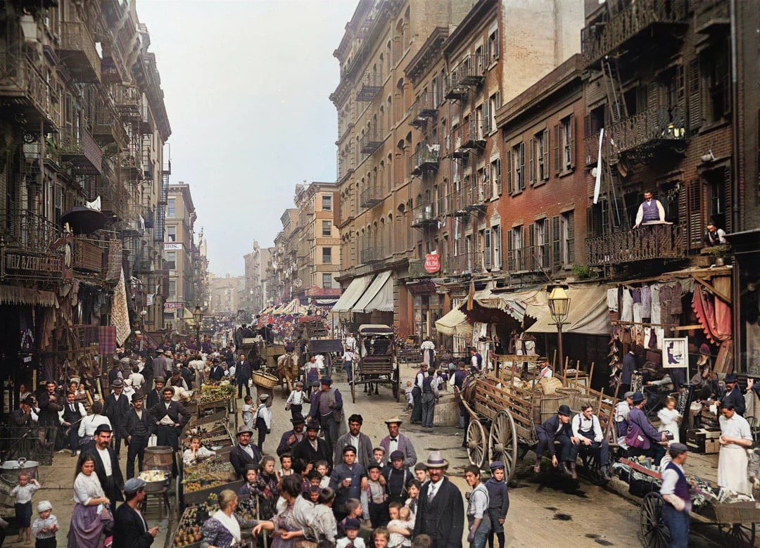 A bustling early 20th-century city street filled with people, market stalls, horse-drawn carts, and tall brick buildings with balconies, showcasing a lively urban neighborhood scene.
