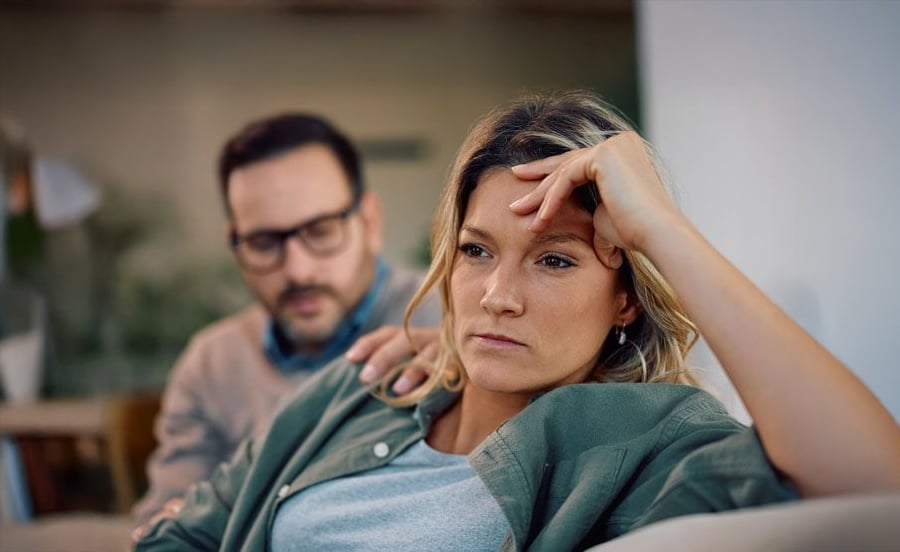 A woman sits on a couch looking distressed, resting her head on her hand, while a man beside her tries to comfort her by putting his hand on her shoulder.