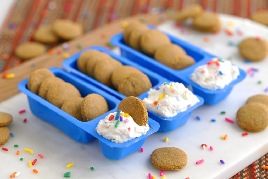 Three blue trays filled with round cookies and whipped cream topped with colorful sprinkles, with extra cookies and sprinkles scattered on a white surface nearby.