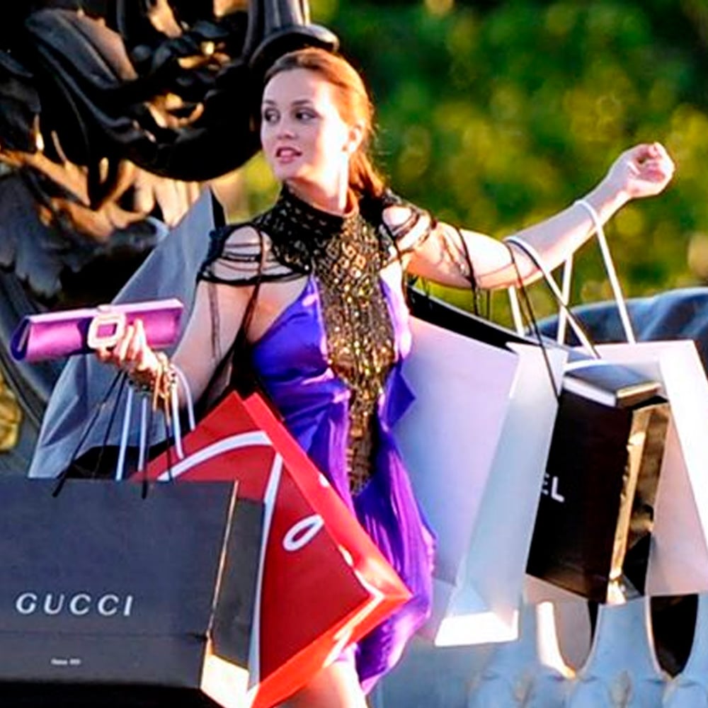 A woman in a purple dress holds several shopping bags from brands like Gucci and Chanel while standing outdoors, looking to the side with her arms full.