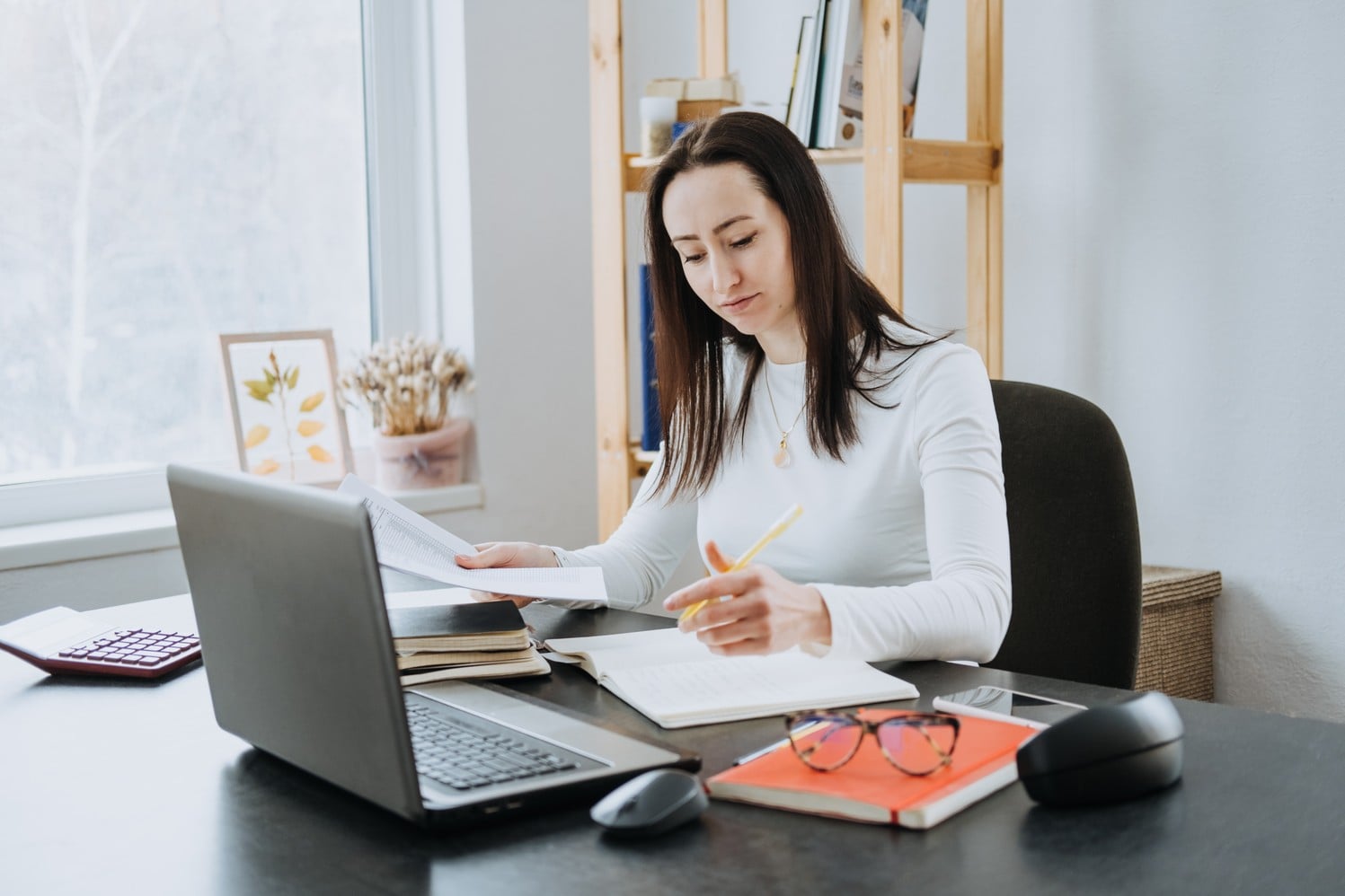 A woman sits at a desk with a laptop, taking notes in a notebook while holding papers. Books, glasses, and a mouse are on the desk. She appears focused, working in a bright, home office setting.