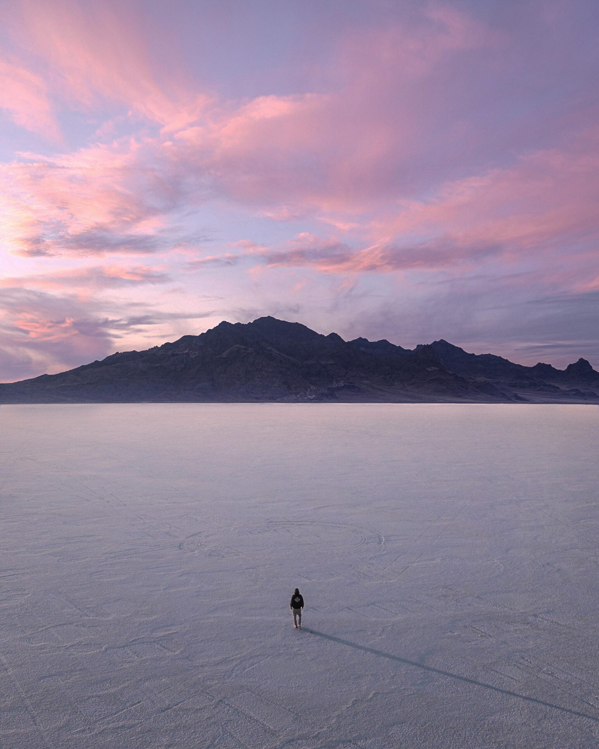 A person stands alone on a vast, flat salt plain at sunset, with a distant mountain range and pink-purple clouds filling the sky.