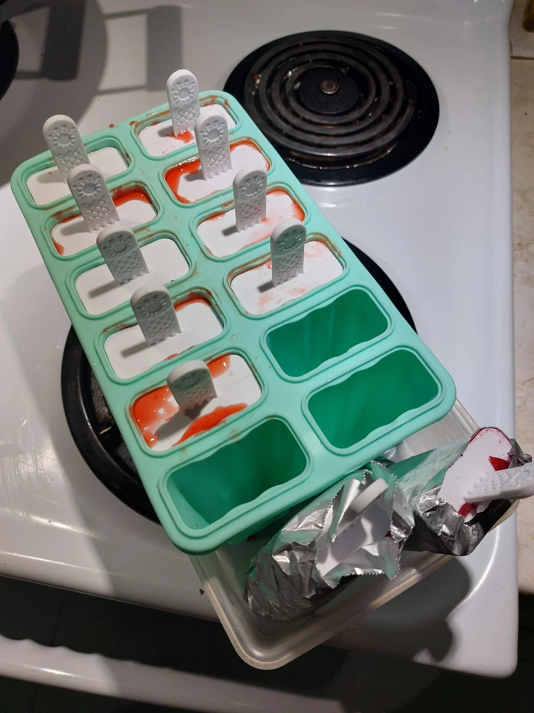 A green silicone popsicle mold with white sticks is filled with a mixture, some spilled red liquid visible, resting on a stove. An open container with foil sits nearby on the stove.