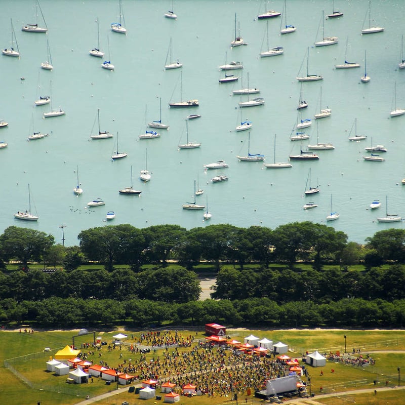 Aerial view of a park event with people gathered around tents on grass, bordered by a line of trees, and many sailboats scattered across calm, light blue water in the background.