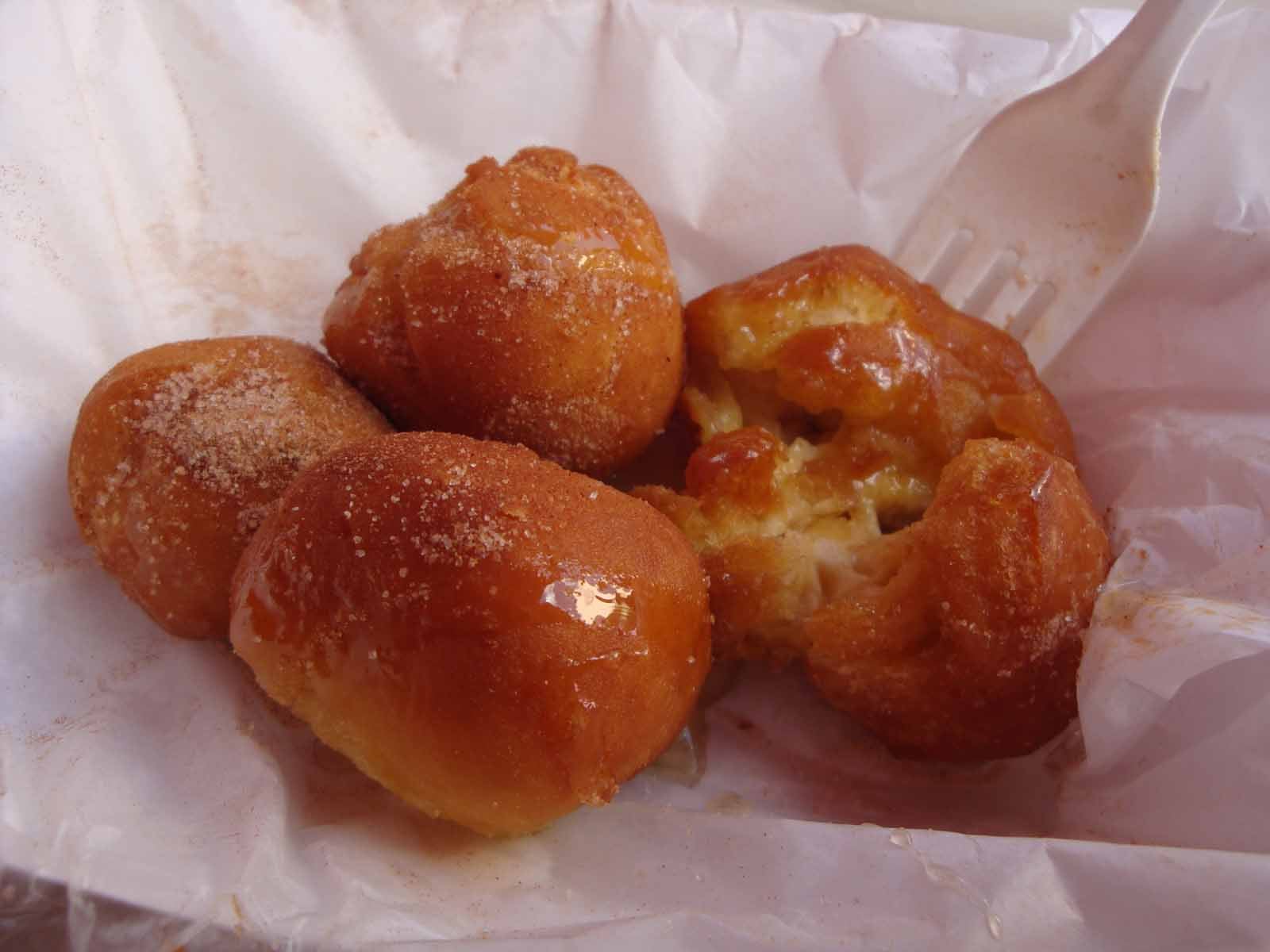 Four golden brown fried dough balls, some covered with sugar and syrup, sit on a piece of white paper. One is partially broken open with a white plastic fork, revealing a soft inside.