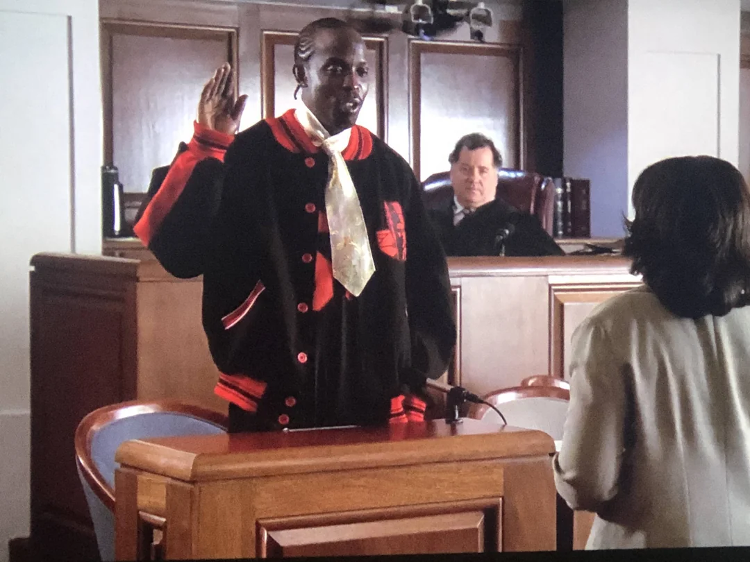 A man wearing a large black and red jacket is being sworn in as a witness in a courtroom, raising his right hand. A judge sits behind the bench, and a woman stands in front of the man.