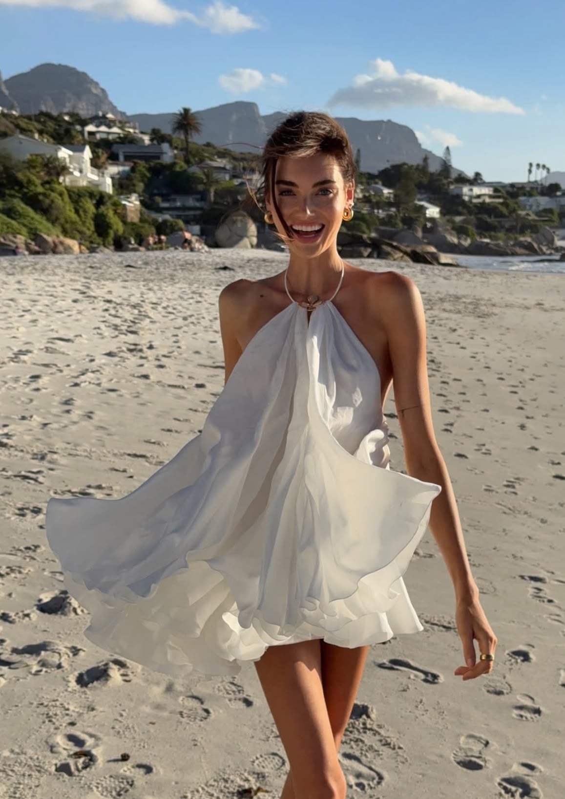 A woman in a flowing white dress smiles while walking on a sandy beach, with coastal houses, green hills, and mountains in the background under a partly cloudy sky.