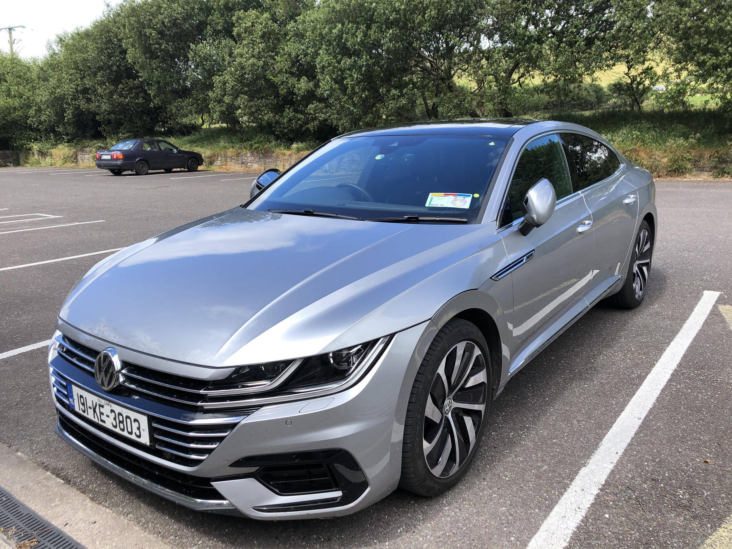 A silver Volkswagen sedan with stylish alloy wheels parked in a nearly empty parking lot, surrounded by green trees and another car in the background.