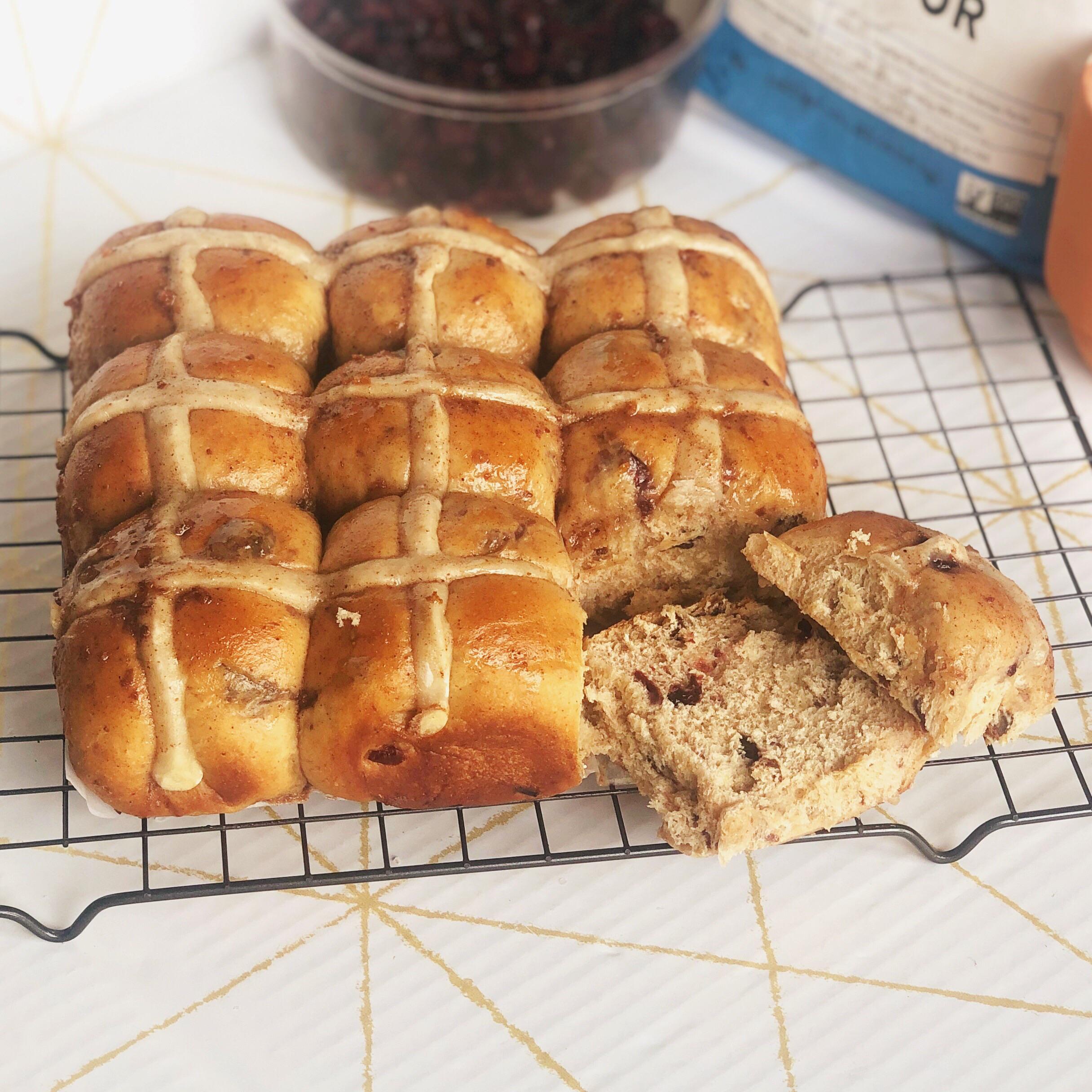 Nine golden brown hot cross buns with white crosses on top are cooling on a wire rack. One bun is split open, showing a fluffy inside with visible dried fruit pieces. Baking ingredients are in the background.