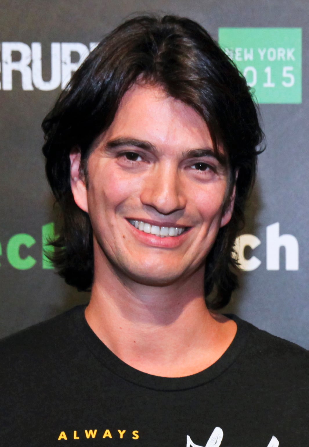 A man with medium-length dark hair and a black t-shirt smiles at the camera against a backdrop with text and logos.