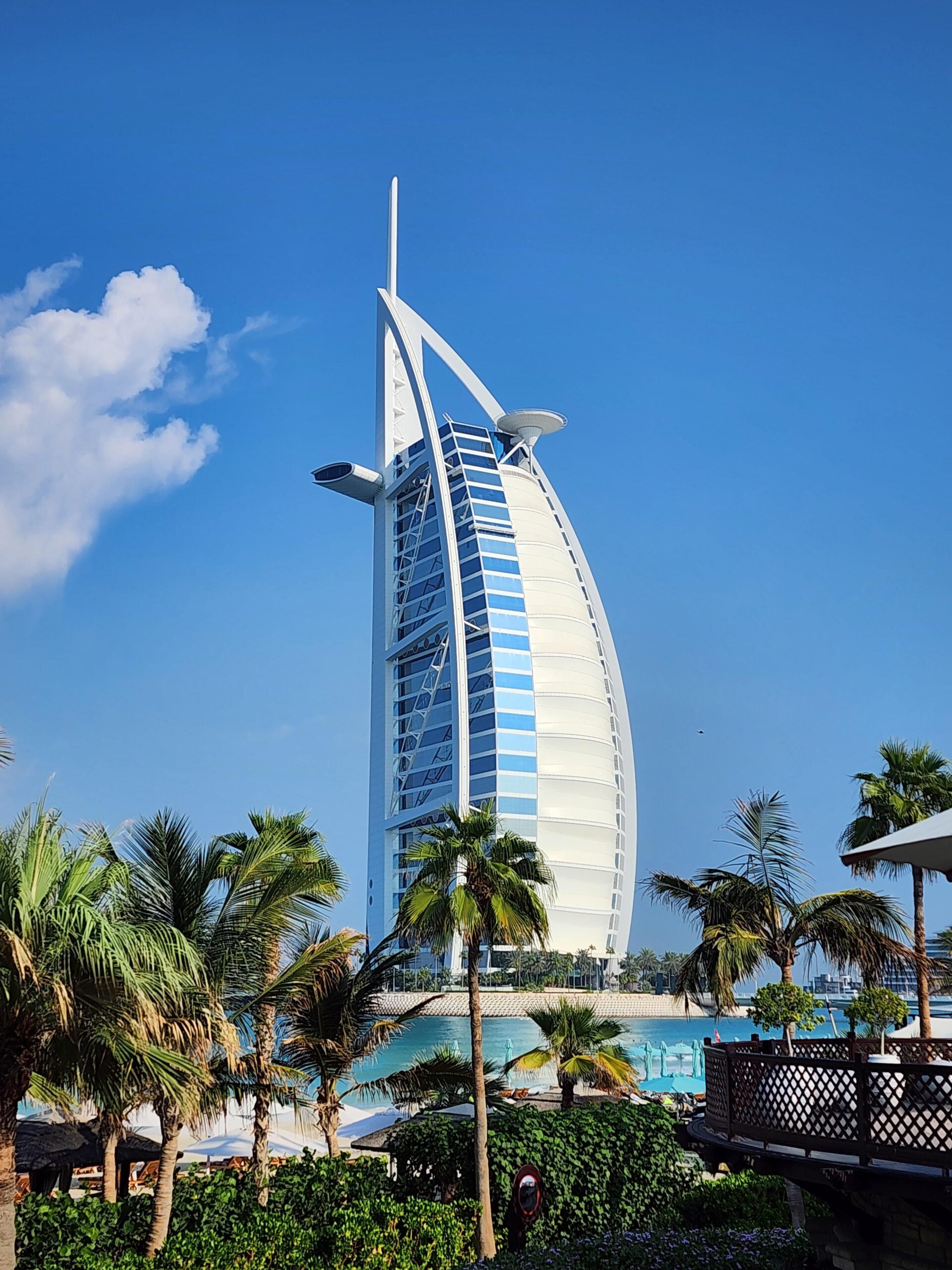 The Burj Al Arab hotel in Dubai stands tall against a clear blue sky, surrounded by palm trees and greenery, with water in the foreground.