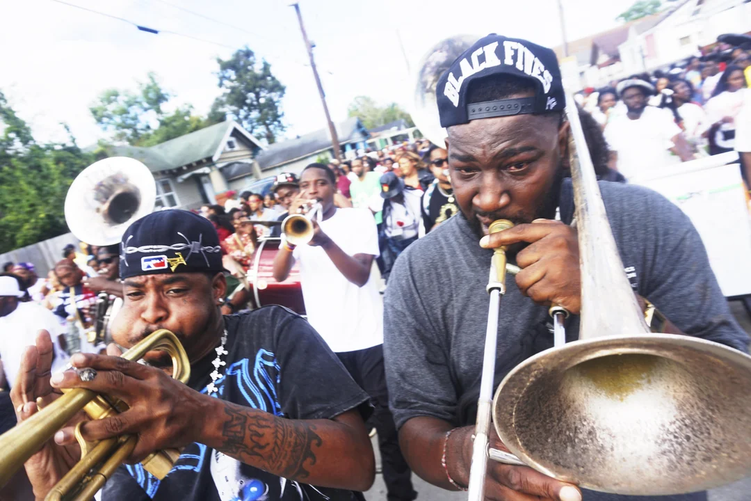 A lively street parade with two men in the foreground playing brass instruments, surrounded by other musicians and people outdoors in a neighborhood setting.