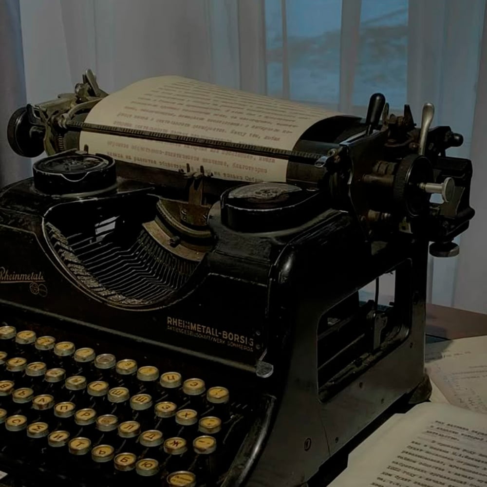 A vintage black typewriter sits on a desk with a typed sheet of paper loaded, surrounded by scattered documents. Sheer curtains and a window are visible in the softly lit background.