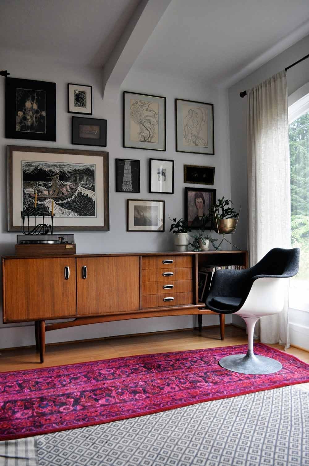 A mid-century modern room with a wooden sideboard, black and white swivel chair, red patterned rug, and a gallery wall of various framed artwork. Sunlight streams through sheer curtains beside a large window.