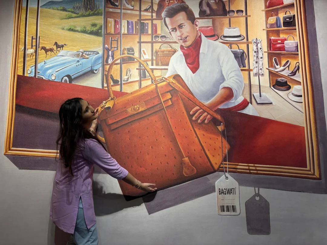 A woman poses with a 3D wall mural showing a man handing her an oversized brown handbag through a picture frame, with a vintage car and hats in the background. A large price tag labeled "BAGWATI" hangs from the bag.