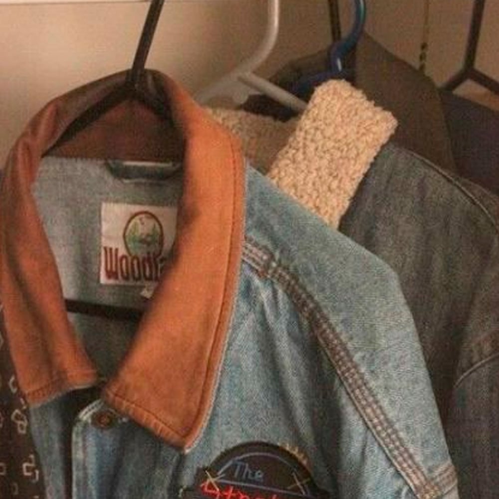 A close-up of denim jackets hanging on hangers in a closet. One jacket has a brown collar and a visible "Woodie" label; another behind it has a cream-colored sherpa lining.