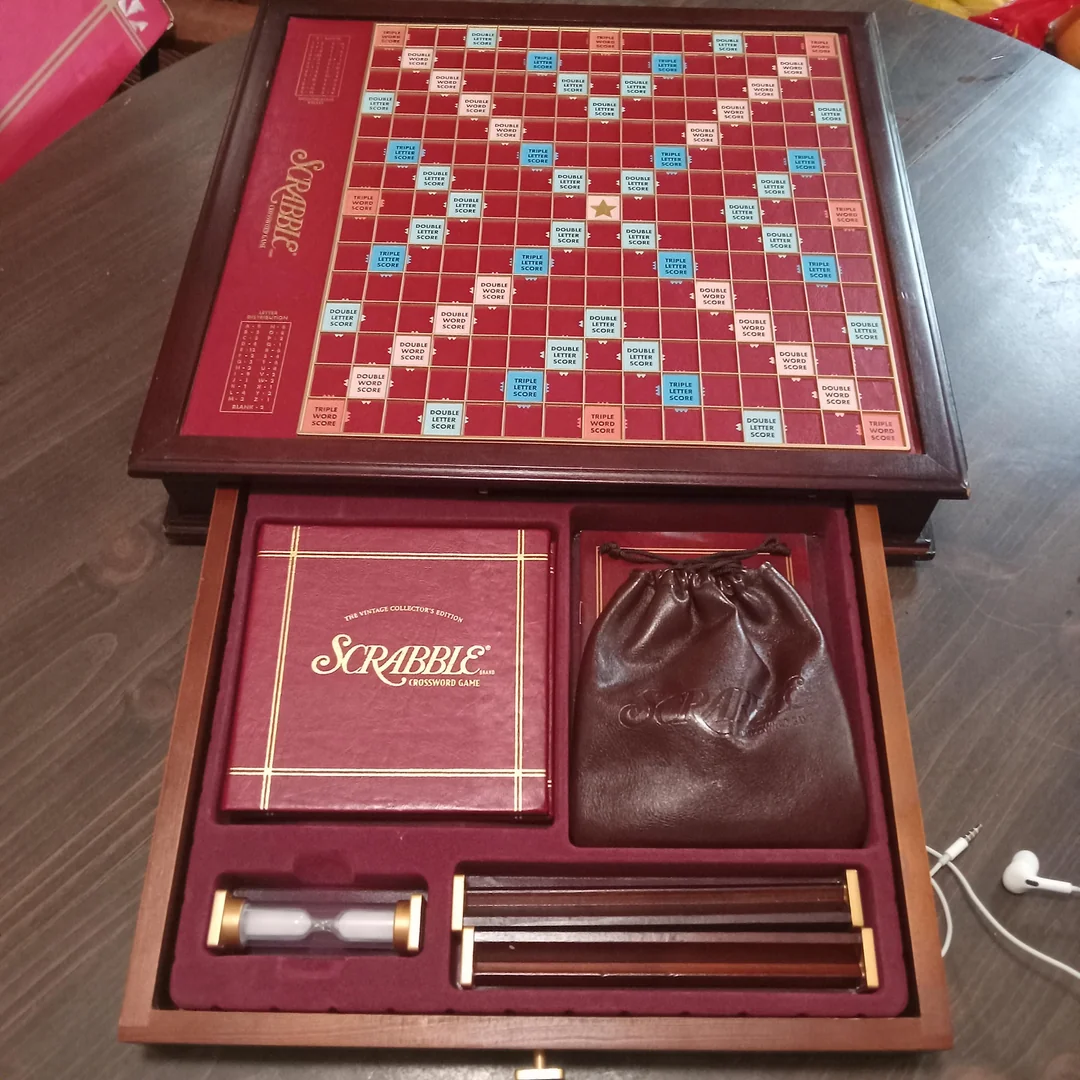 A deluxe Scrabble board game set with a wooden board, tile racks, score pad, timer, and a brown bag for tiles, all neatly stored in a wooden box with compartments.