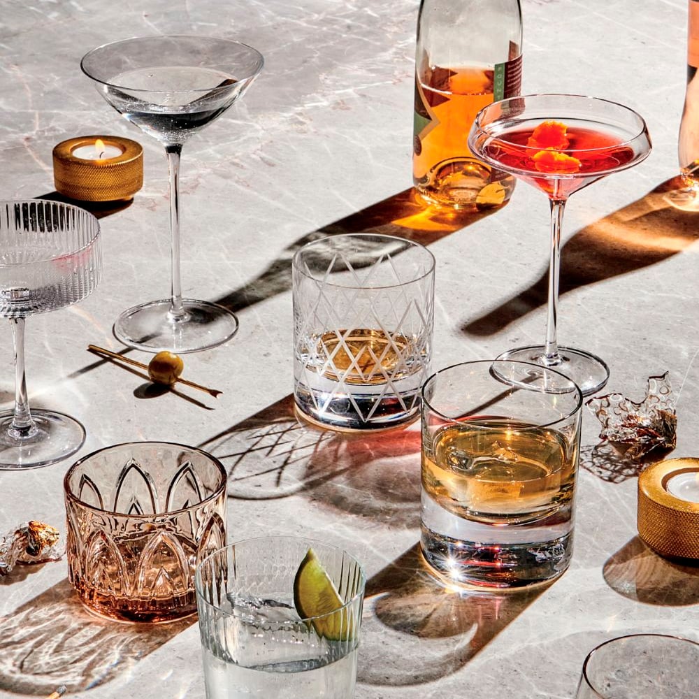 A variety of elegant cocktails and spirits in different glassware are arranged on a marble surface, with candles and garnishes like olives, orange slices, and a lime wedge. Shadows are cast across the scene.