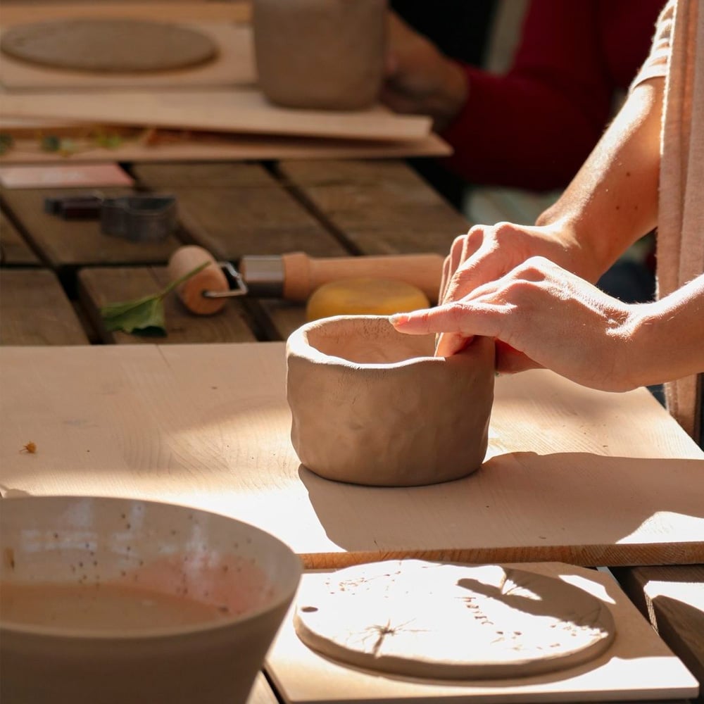 A person shapes a small clay pot with their hands at a wooden table, surrounded by pottery tools and other clay items, in a sunlit pottery workshop.