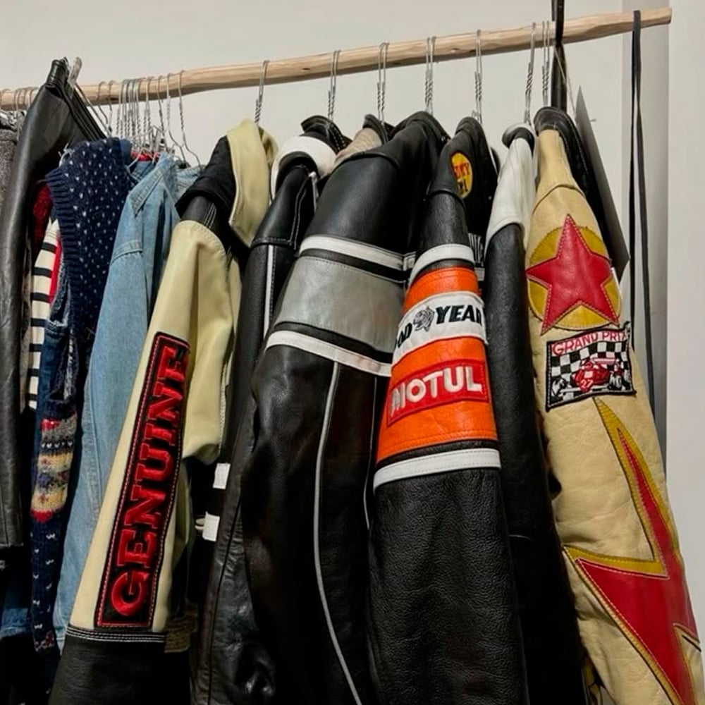 A clothing rack with various vintage leather jackets, many featuring bold patches and logos, such as "GENUINE," "MOTUL," racing badges, and a large red star on a cream background.