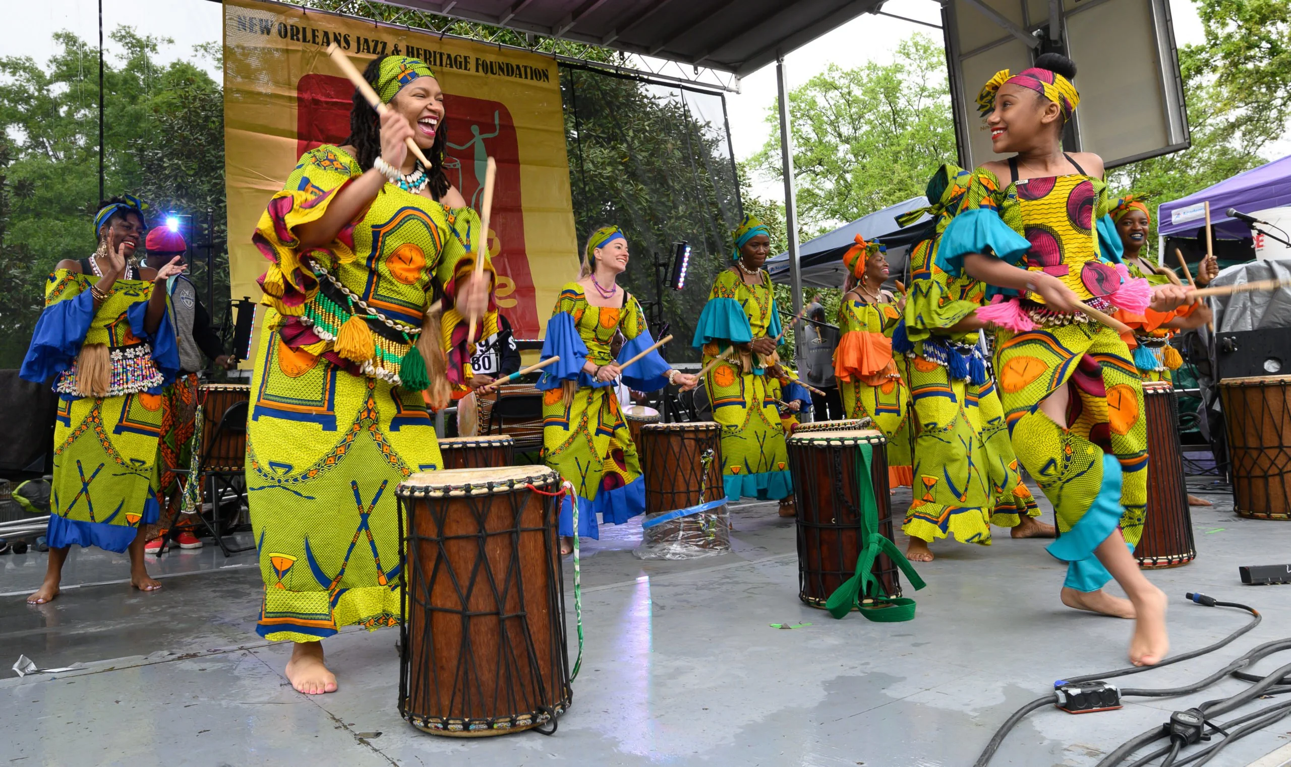 A group of women in colorful, patterned outfits joyfully dance and play drums onstage at an outdoor festival, with a New Orleans Jazz & Heritage Foundation banner in the background.