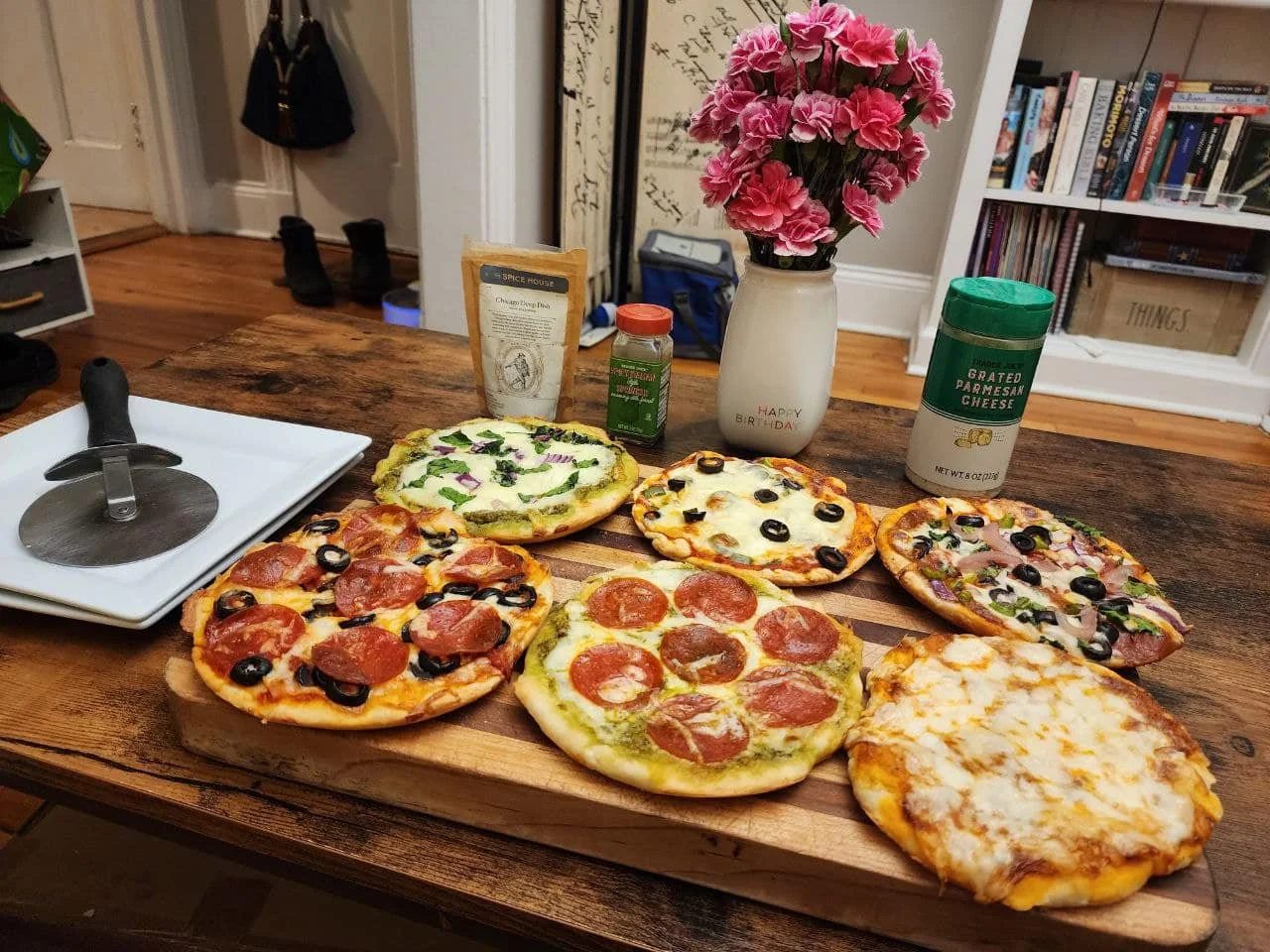 A wooden table with six homemade pizzas featuring various toppings like pepperoni, olives, and vegetables. Nearby are a pizza cutter, grated cheese, Italian seasoning, a vase of pink flowers, and kitchen shelves in the background.