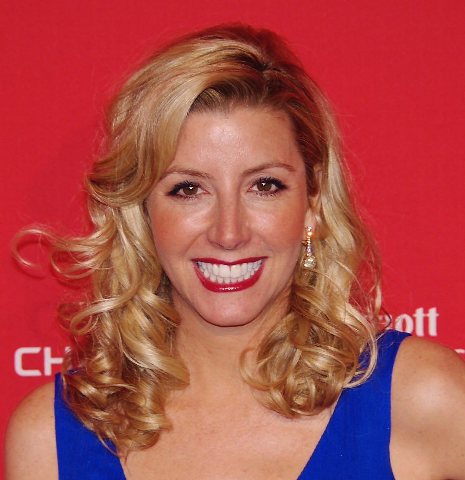 A woman with wavy blonde hair, wearing a blue dress and dangling earrings, smiles in front of a bright red background.