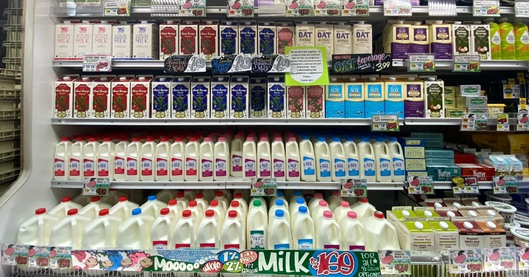 Shelves in a grocery store filled with various milk cartons and bottles, including whole, 2%, and flavored milk options, arranged in rows with colorful price and product labels displayed below them.