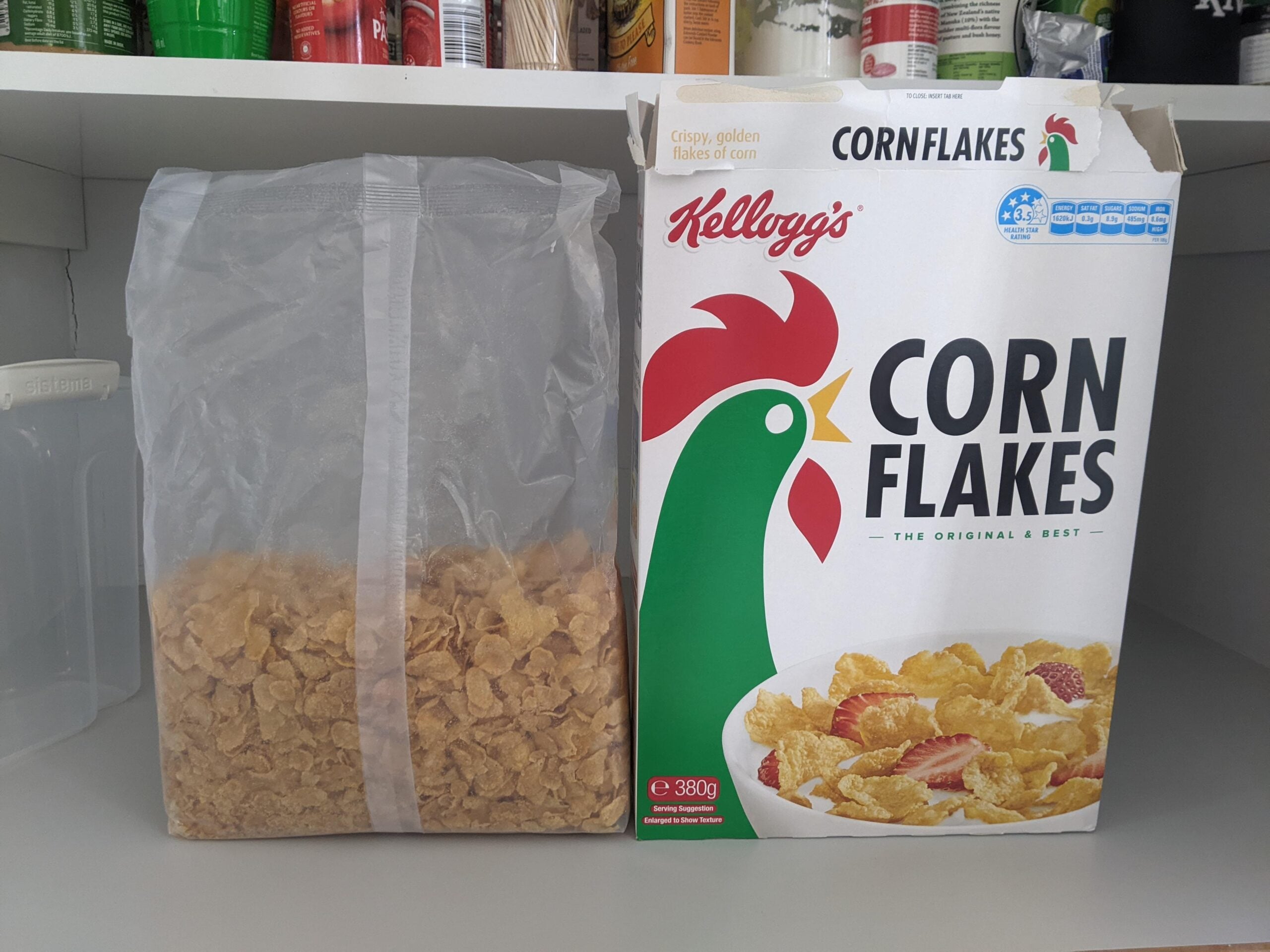 A box of Kellogg’s Corn Flakes stands open on a shelf next to a clear plastic bag filled with corn flakes cereal. The box features a green and red rooster and a bowl of cereal with strawberries.