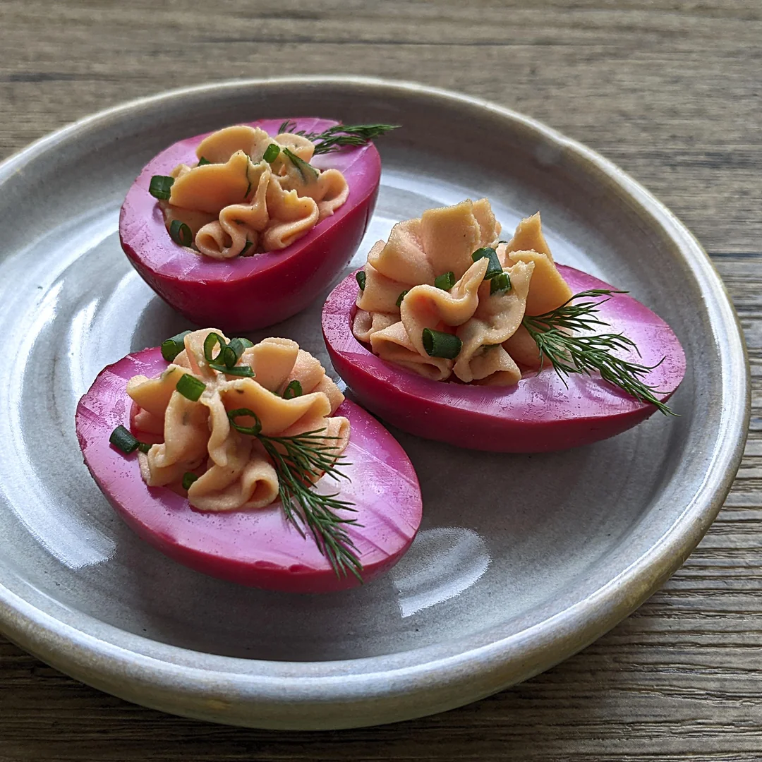 Three halved pink pickled eggs on a gray plate, each filled with a whipped filling and garnished with fresh dill and chopped chives, set on a wooden table.